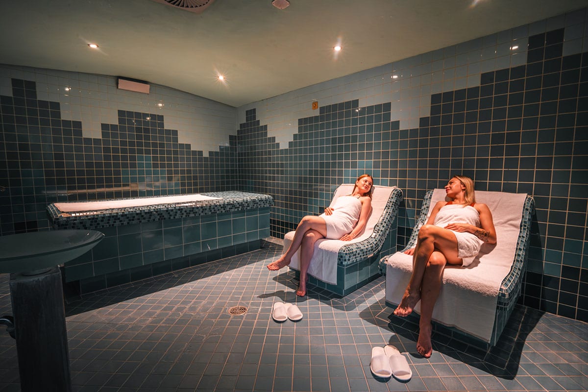 Two women in towels relaxing on heated loungers in a steam bath